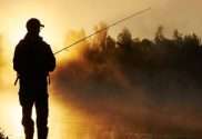 A man silhouetted on the water holding a long fishing pole as the sun sets in the background in a forest.