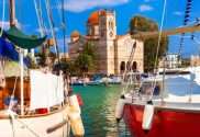Red and beige sailboats sitting next to one another in a marina in front of a building beneath a blue sky.
