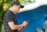 A man wearing a black baseball cap and shirt standing in front of a boat with a clipboard while taking notes.