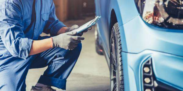 Smaller Things You Shouldn’t Overlook in Your Car 1 A mechanic kneels on one knee, holds a clipboard, and inspects the blue car and tires sitting in front of him.
