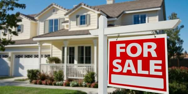 A cream-colored, two-story house features a grassy lawn, trees, and a "for sale" sign in front of it.