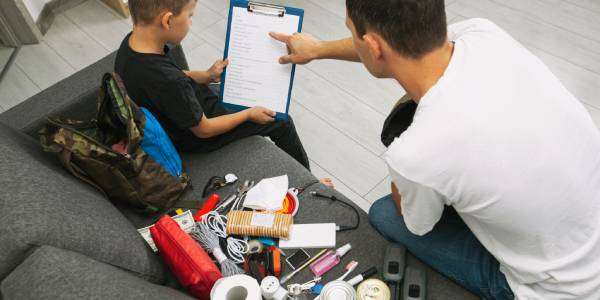 3 Emergency Survival Supplies To Keep at Home 1 A man points to a checklist a young boy is holding. They sit on a couch surrounded by emergency supplies.