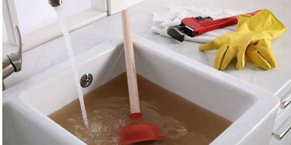 Home And Remodeling Expo is This Sat & Sun - Feb 8th & 9th 42 A kitchen sink with the faucet on is filled with brown water, signifying a clog. A plunger and other tools are nearby.