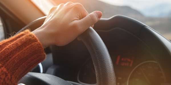 A close-up view shows a hand resting on a black steering wheel of a vehicle. The dashboard is behind the steering wheel.