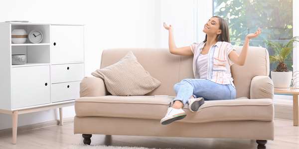 5 Tips for Improving Your Home’s Indoor Air Quality 2 A woman sits on a tan couch with her eyes closed, head tilted back, and arms reaching up. Sunshine comes through the window.