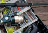 A tackle box lies open on a wooden deck. Many lures and other tools are inside with a rod laying on top of the box.