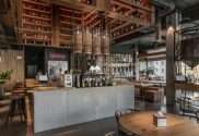 The interior of a loft style restaurant with a bar in the center. It uses wooden surfaces, bar seating, and hanging lights.