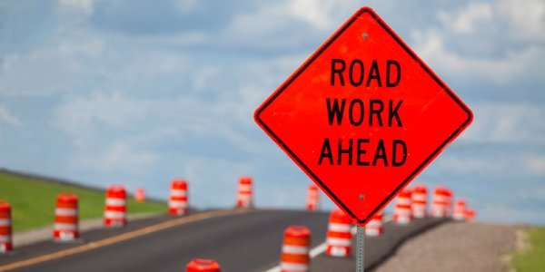 An orange sign saying road work ahead in front of a road that's under repair. Orange barrels are lined up on the road.